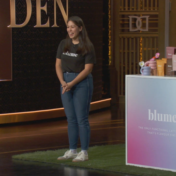 A person stands smiling on a stage next to a product display with blume written on it. Theyre wearing a black blume T-shirt and jeans. The background has a large illuminated sign with DEN visible.